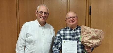 Zwei Männer mittleren Alters stehen nebeneinander; der Mann rechts im Bild trägt ein blaukariertes Hemd und hält einen Blumenstrauß und eine Urkunde in der Hand; der Mann links im Bild ist der Bürgermeister; er trägt ein weißes Hemd und gratuliert dem anderen.