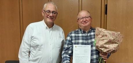 Zwei Männer mittleren Alters stehen nebeneinander; der Mann rechts im Bild trägt ein blaukariertes Hemd und hält einen Blumenstrauß und eine Urkunde in der Hand; der Mann links im Bild ist der Bürgermeister; er trägt ein weißes Hemd und gratuliert dem anderen.