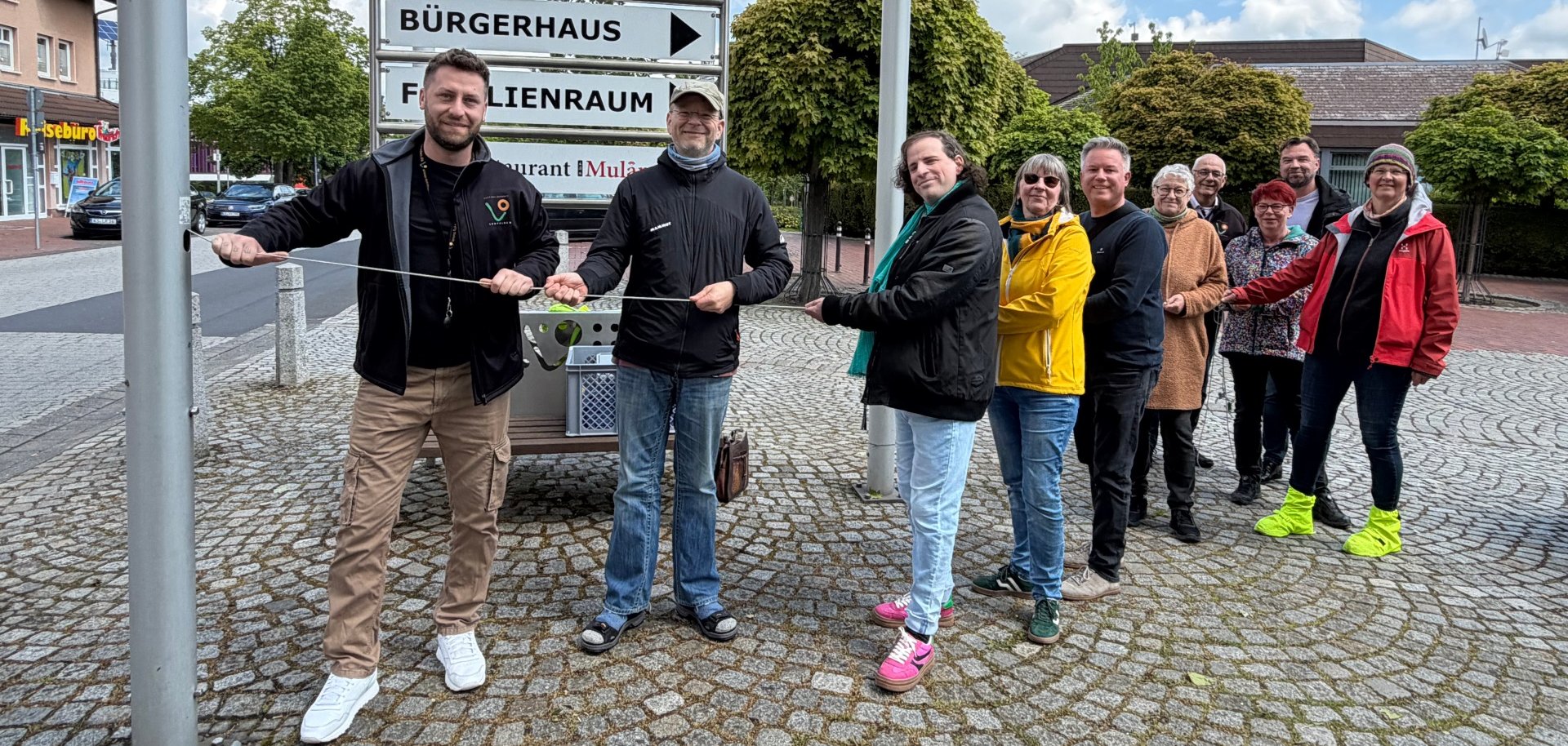Mehrere Personen ziehen am Seil, das die Regenbogenfahne am Fahnenmast nach oben bringt.