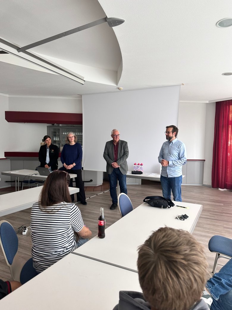 Begrüßung im Familienraum durch Bürgermeister Uwe Jäger (2.v.r.), Klimaschutzmanagerin Ursula Paul (2.v.l.), Radverkehrsbeauftragte der Gemeinde, Angelina Michel (l.), und Radverkehrsbeauftragter der Söhre-Schule, Simon Dölle (r.). Zwei Frauen und zwei Männer in einem Raum mit Holzfußboden for einer weißen Leinwand.