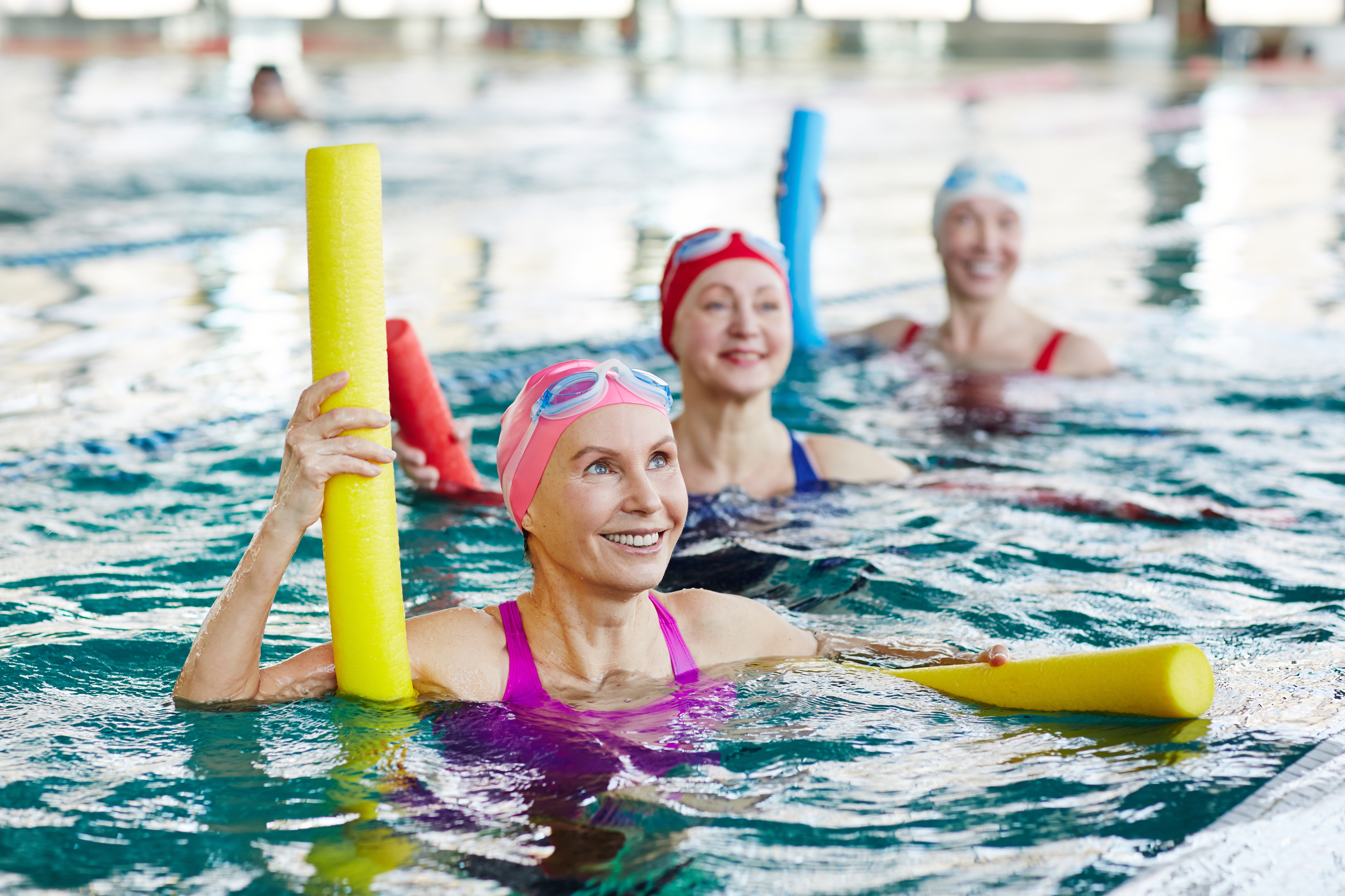 Lächelnde Frau in Badekleidung und zwei weitere aktive Frauen, die den Ratschlägen des Trainers bei der Gymnastik im Wasser zuhören.