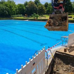 Auf dem Foto sieht man einen Teil des Schwimmbeckens des Freibads Lohfelden sowie eine Absperrung mit rot-weißem Flatterband und Teile einer Baggerschaufen.