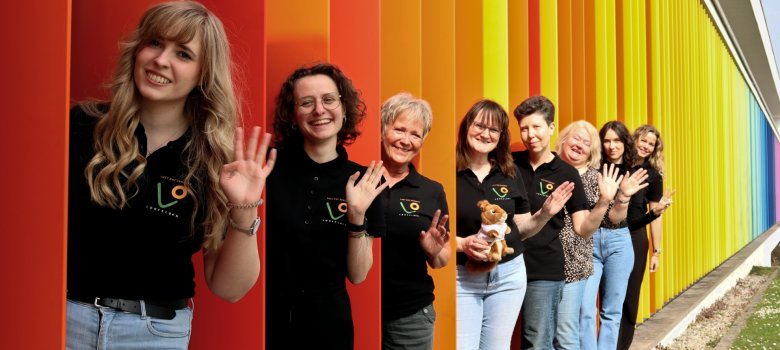 Gemeinde- und Schulbücherei Lohfelden Team der Bücherei steht hintereinander in den bunten Lamellen; winkt.