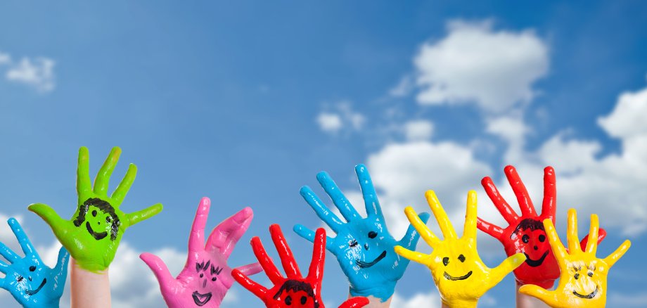 Auf dem Foto sind acht Kinderhände zu sehen, die in bunten Farben bemalt sind und sich in einen blauen Himmel mit weißen Wolken erstrecken. 