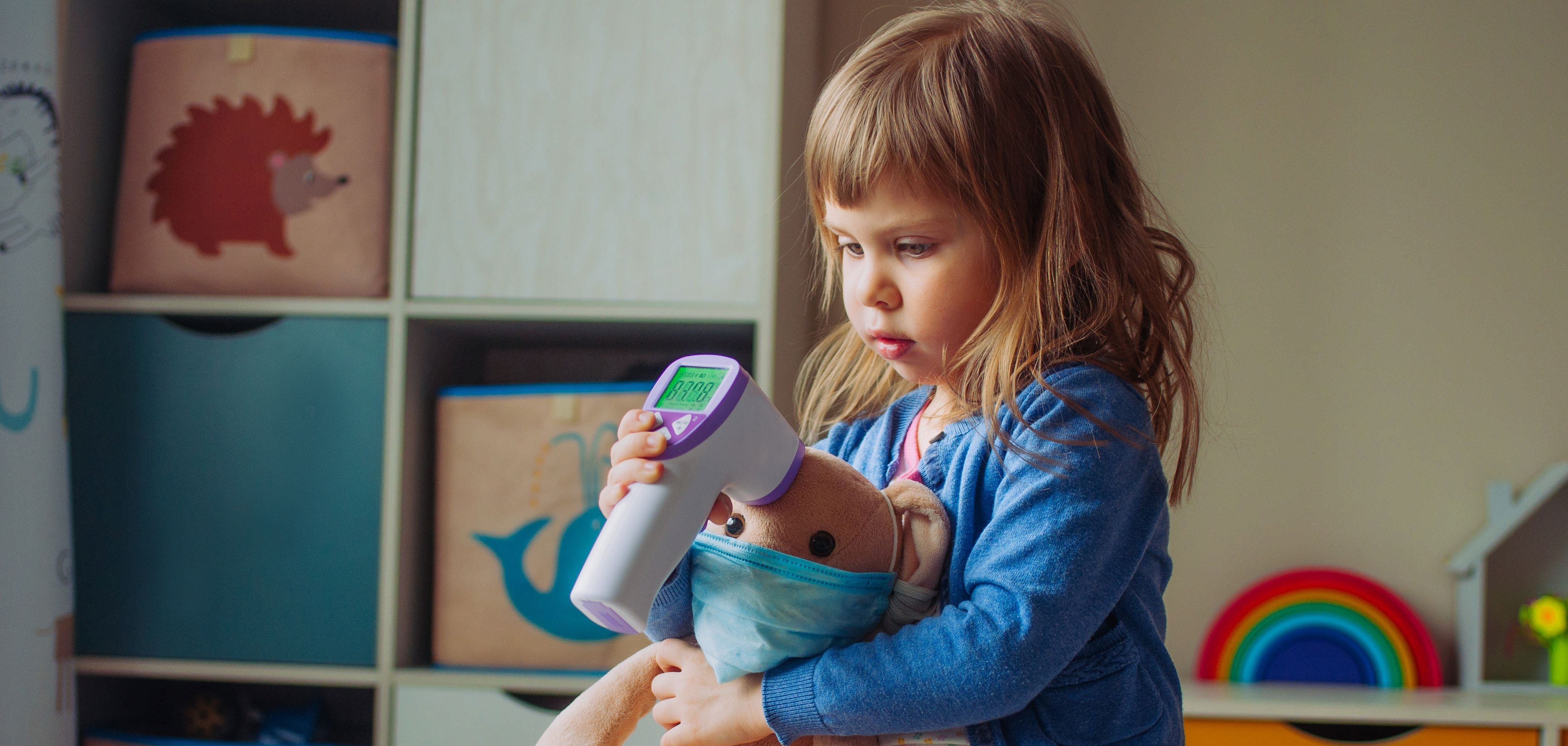 Auf dem Foto sieht man ein kleines Mädchen mit halblangen mittelblonden Haaren mit Pony, das bei einem kleinen Stoff-Teddy Fieber misst.
