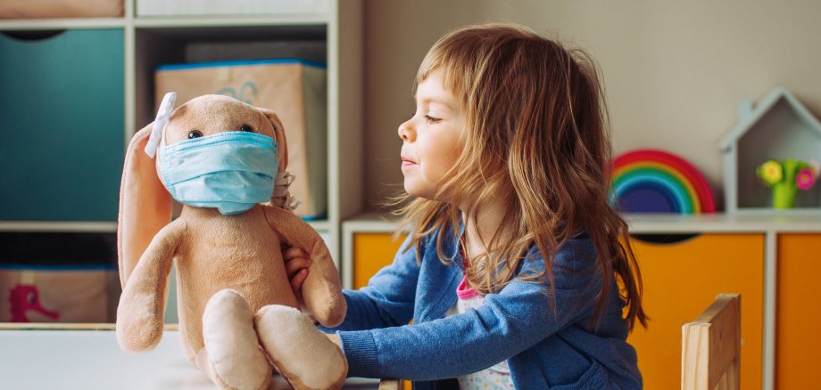 Kleines Mädchen sitzt in der Kindertagesstätte und spielt mit einem Kaninchen-Plüschtier, das eine medezinischen OP-Maske trägt trägt.