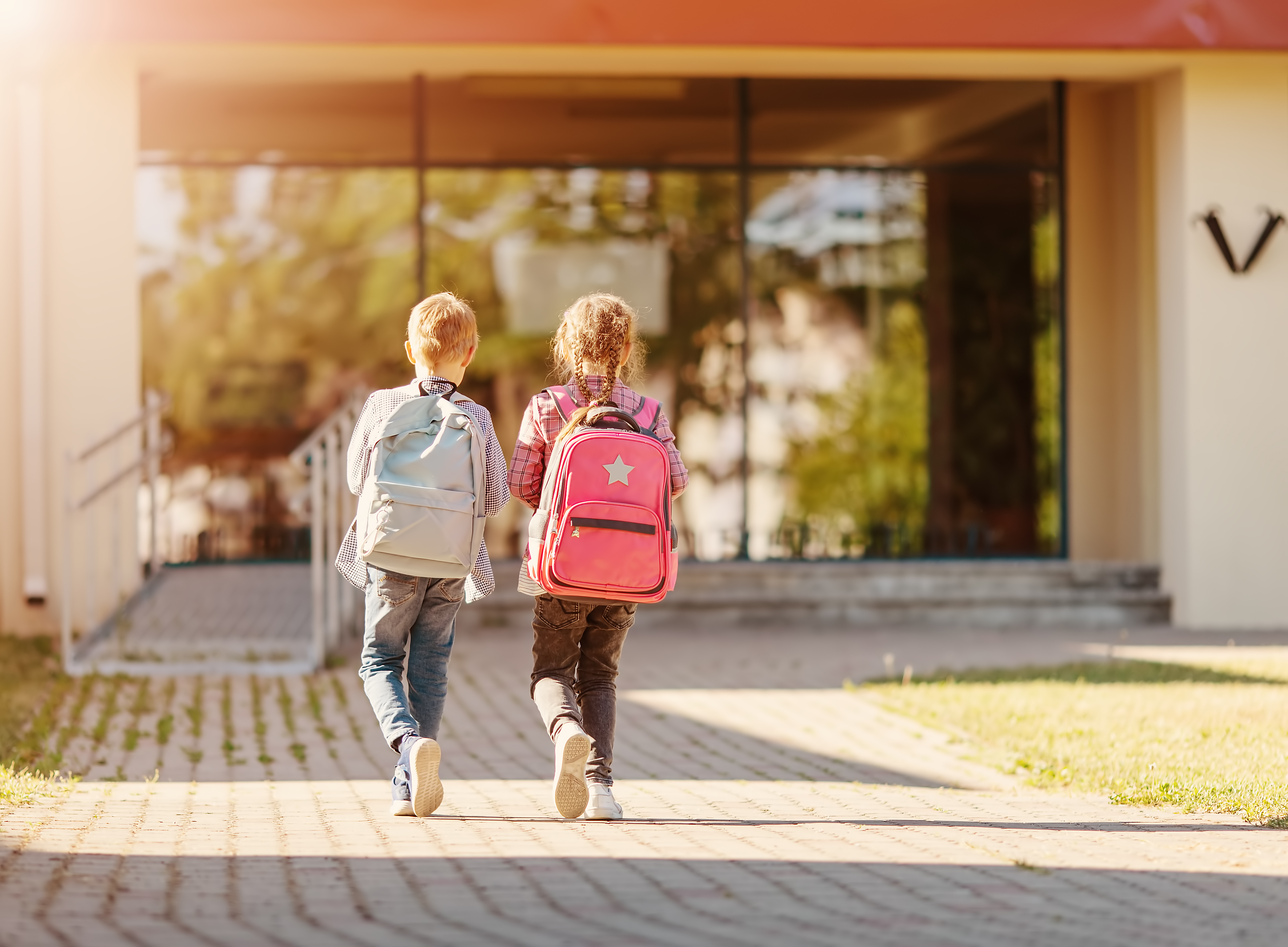 Mädchen und Junge mit Schulranzen gehen zusammen zur Schule, Foto von hinten.