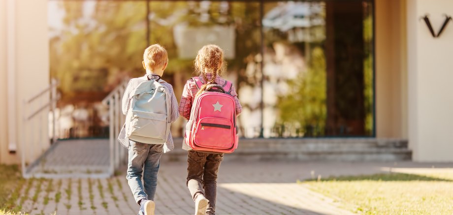 Mädchen und Junge mit Schulranzen gehen zusammen zur Schule, Foto von hinten.