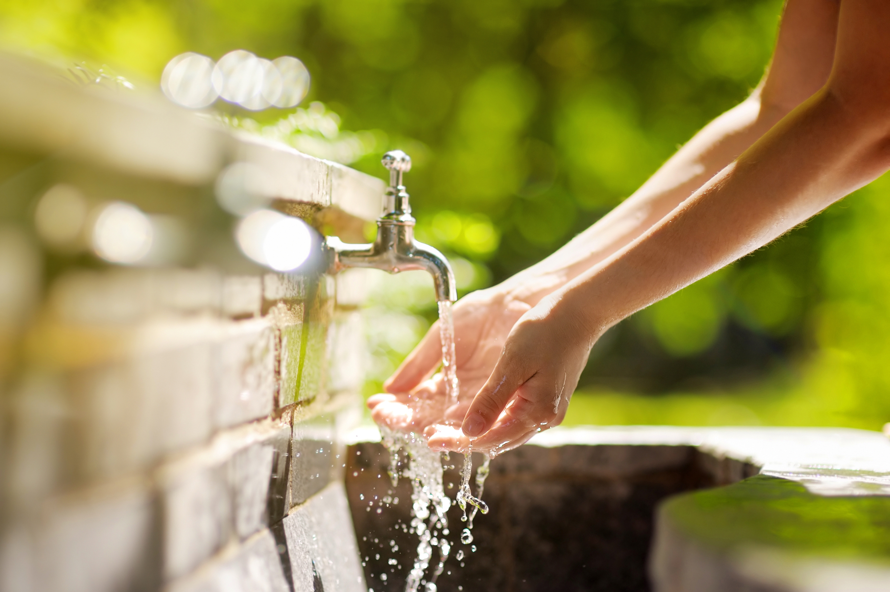 Auf dem Foto sieht man vor grünem Hintergrund zwei Hände unter einem geöffenten Wasserhahn, aus dem klares Wasser sprudelt.
