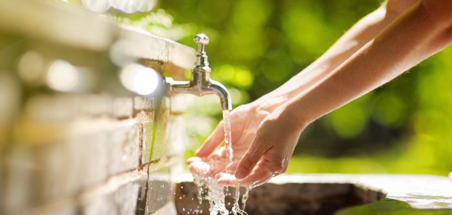 Auf dem Foto sieht man vor grünem Hintergrund zwei Hände unter einem geöffenten Wasserhahn, aus dem klares Wasser sprudelt.