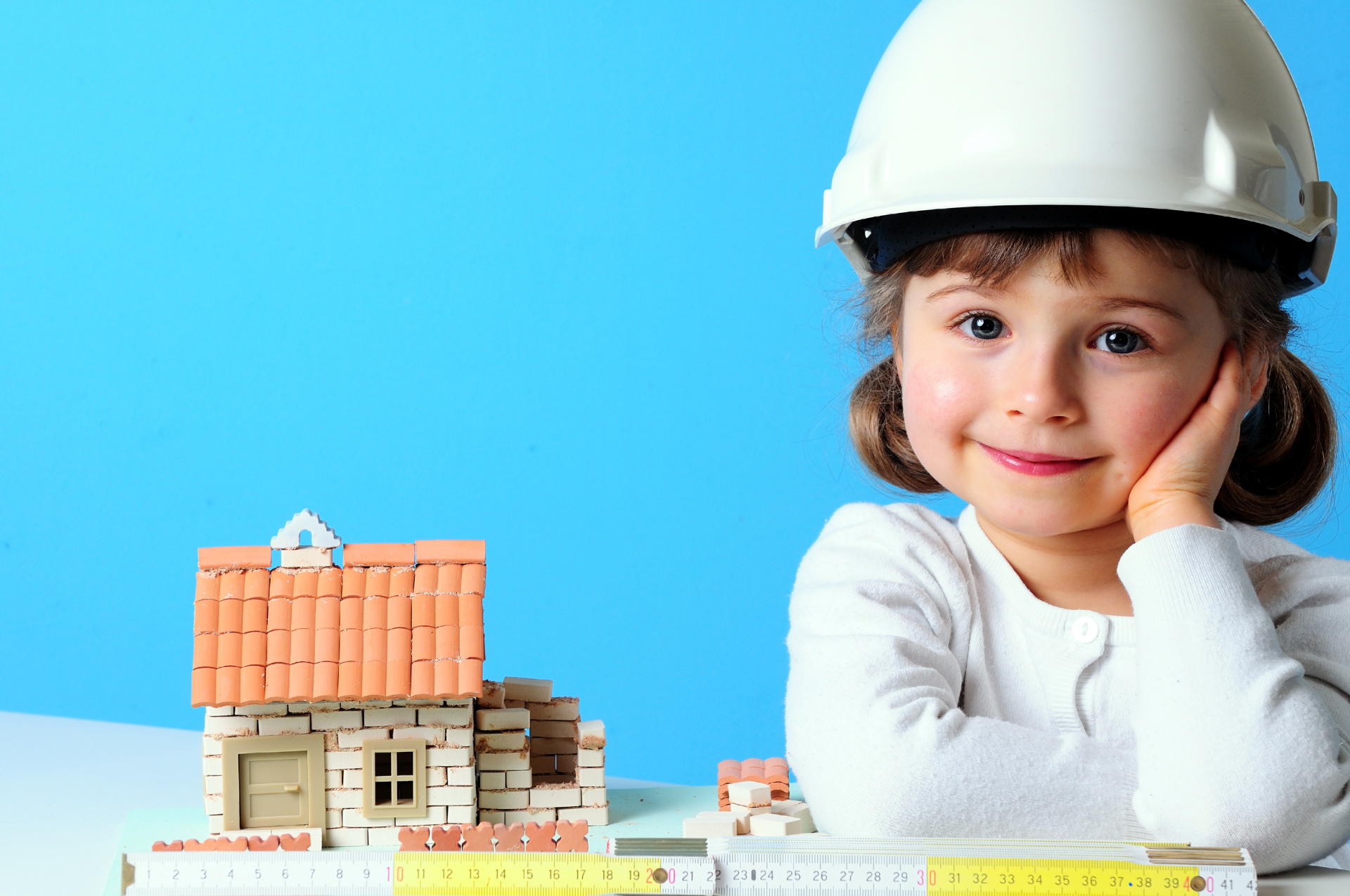 Klimaschutz in Lohfelden - Wärmeplanung Kleine Bauarbeiterin – Hübsches Mädchen mit weißem Bauhelm neben einem kleinen Haus im Bau vor blauem Hinterbrund