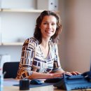 Auf dem Foto ist eine lächelnde junge Frau zu sehen, die in einem Büro sitzt und am Computer arbeitet.