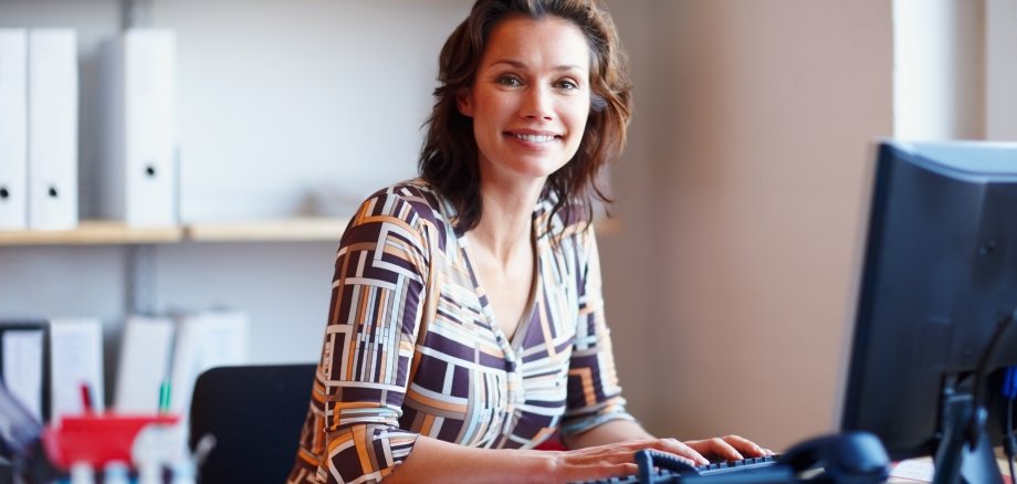 Auf dem Foto ist eine lächelnde junge Frau zu sehen, die in einem Büro sitzt und am Computer arbeitet.