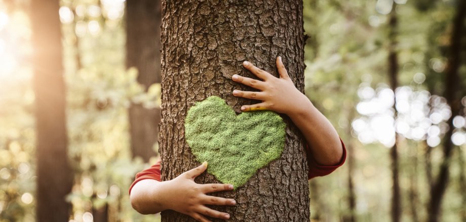 Natur ... ein Kind umarmt einen Baum, auf dem ein Moos-Herz zu sehen ist.